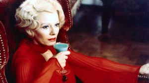 Delphine Seyrg assise en robe rouge, porte un verre à ses lèvres dans le film Les lèvres rouges.