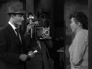 Un journaliste mesquin interroge dans la rue une femme apeurée témoin d'un crime, entre eux deux, un lourd appareil photo des années 50, sur pied, scène du film L'inexorable enquête.