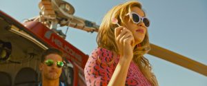 Une jeune femme blonde tient une sucette à la main, un look années 60 avec lunettes de soleil rondes et chemisier rose, derrière elle un jeune homme aux lunettes aviateur, et le sommet d'un hélicoptère, plan du film Revenge de Coralie Fargeat.