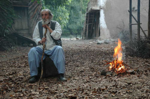 Un vieillard assis, canne sous le menton, près d'un feu de bois, scène du film L'enclave.