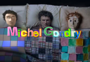 Michel Gondry est allongé dans un lit entre deux marionettes à son effigie, l'une en carton, l'autre en papier, pour notre article analyse de sa filmographie.