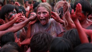 Une jeune femme blonde hurle au milieu des cannibales à la peau rouge qui lui touchent les cheveux., scène du film The Green Inferno.
