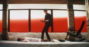 Un yakuza,  l'arme au poing, est debout sur ses gardes près du cadavre d'une femme en robe blanche, sur fond rouge et blanc, plan stylisé conçu par Seijun Suzuki dans un de ses films.