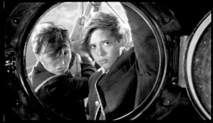 En noir et blanc, Vimala Pons et une autre jeune femme, vêtus à la garçonne sont sur un bateau, les cheveux au vent, la mine soucieuse ; nous les voyons à travers un hublot ; scène du film Les garçons sauvages.