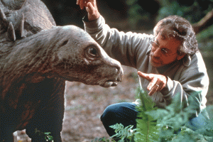 Steven Spielberg prépare son plan accroupi tout près d'une maquette de dinosaure sur le tournage de Jurassic Park.
