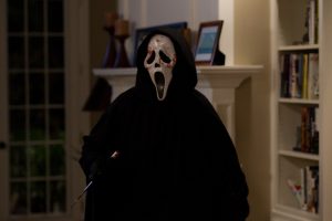 Le tueur Ghostface se tient debout dans un salon, du sang sur son masque, et brandit son couteau, scène du film Scream.