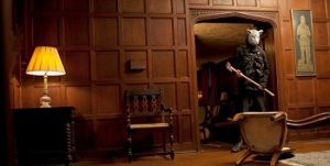 Un homme avec un masque de lapin tient une massue dans la main se tient sur le palier de la porte d'un grand salon tout en bois, scène du film You're next pour notre analyse du home invasion.