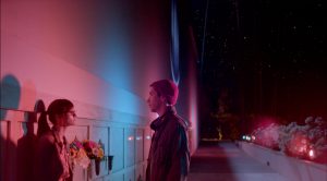 Un jeune homme et une jeune femme face à face dans une rue déformée par l'angle de prise de vue, et éclairée de néons bleus et roses, scène du film Comet.