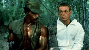 Jean-Claude Van Damne sur le tournage du film Predator ayant une prothèse de bras Predator sur un bras, à côté de lui, l'acteur Carl Weathers en tenue de soldat, en fond, la jungle amazonienne.