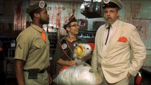Trois hommes dans la cuisine du fast-food avec du sang sur les murs, deux employés dont l'un regarde leur chef avec admiration, le chef avec les mains dans les poches et le regard au loin, scène du film Poultrygeist.