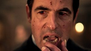 Le Comte Dracula, les yeux injectés de sang, passe le doigt sur ses lèvres, comme s'il regardait une future proie, scène de la mini-série Netflix Dracula.