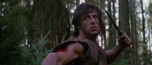 John Rambo le couteau à la main, apeuré, s'apprête à frapper quelqu'un dans la forêt.