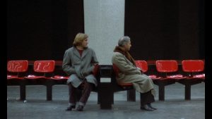 Gérard Depardieu sur les sièges rouges du quai du RER à Paris, tourne la tête vers l'homme assis à côté de lui qui lui tourne le dos, scène du film Buffet Froid.