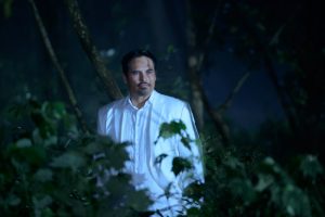 L'acteur Michael Pena dans un beau costume blanc, l'air serein et les mains dans les poches, debout derrière les feuillages d'une forêt de nuit, scène du film Nightmare Island.
