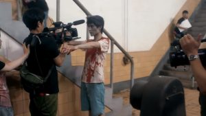 Un zombie au t-shirt blanc maculé de sang se tient devant un cadreur les bras devant, scène de tournage dans le film Ne coupez pas !