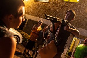 Dans une rue de nuit, , éclairée par une lumière aux teintes jaunes, un homme menace un autre avec un fusil à pompe, derrière eux, un jeune homme en débardeur jaune et une adolescente, scène du film Mega Time Squad. 