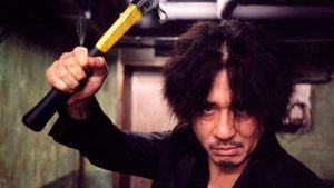 L'acteur Choi Min-Sik brandit son marteau jaune et noir, face caméra, scène du film Old Boy.