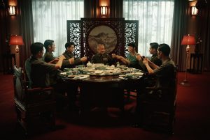 Une tablée d'hommes portent un toast, devant un sompteux paravent asiatique, scène du film Nina Wu.