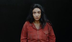 Sur fond noir, l'actrice Souhir Ben Amara nous regarde, l'air ahuri, portant une chemise rouge sale, scène du film Sortilège.
