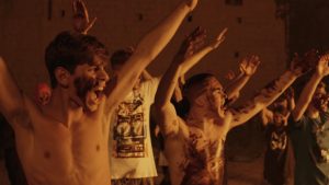 Les jeunes hommes sauvages du film Piranhas hurlent les bras levés, face à un feu, au premier plan d'entre eux sont torse nu et portent des traces de boue sur le visage et la poitrine.