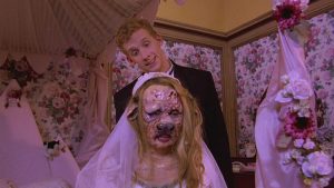 Un des invités du mariage tout sourire derrière une Juliet monstrueuse à groin de cochon, en robe de mariée, scène du film Tromeo et Juliet.
