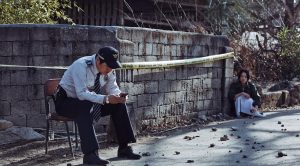 Le héros policier du film The Strangers patiente assis sur une chaise dans la rue sous le soleil, à quelques mètres près de lui, le fixe un jeune vagabond inquiétant.