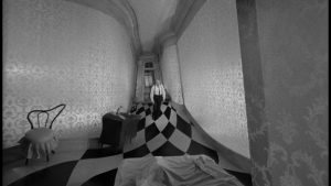 Vision subjective complètement déformé d'un couloir au sol en damier, au premier plan le pied d'un lit, au fond de l'image un homme de face qui s'approche, scène du film Seconds.