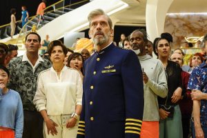 Hugh Laurie en capitaine, uniforme bleu, au premier plan, derrière lui tout l'équipage : ils regardent tous au loin apeurés, scène de la série Avenue 5.
