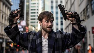 Daniel Radcliffe, vêtu et barbu comme un mendiant, lève les mains en l'air comme pour se rendre en pleine rue, dans ses poings, deux revolvers, scène du film Guns Akimbo.