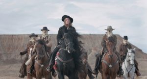 Cinq malfrats à cheval devant une montagne, tous les visages sont encagoulés sauf celui de leur chef, une femme, en tête sur son cheval noir, scène du film L'etat sauvage.