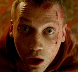 Andrei les yeux exorbités, face caméra, du sang sur la tempe gauche, en fond un tapis en moquette aux motifs viellots, scène du film Why don't you just die.