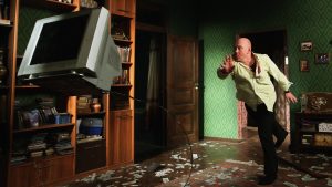 Un homme chauve au milieu d'un salon lance une vieille télévision cathodique, scène du film Why don't you just die.