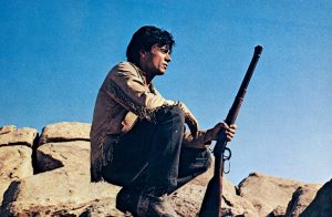 Sur un fond de ciel bleu sans nuage et de rochers arides, un jeune homme est accroupi, le fusil dans la main crosse contre terre, et regarde devant lui, l'horizon à sa gauche, scène du film Willie Boy.