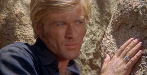 Portrait de Robert Redford dans le film Willie Boy, la main contre un rocher et le regard tourné vers la gauche sous un soleil de plomb.