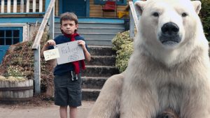 Un petit garçon à l'écharpe rouge montre deux dessins sur le palier de sa maison, à côté de lui un ours blanc, scène du film Timmy Failure, des erreurs ont été commises.