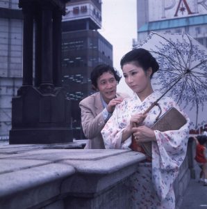 Une geisha, parapluie ouvert dans la rue, est accostée par un homme qui lui pose sa main sur son épaule de derrière elle.