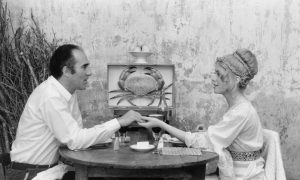 Michel Piccoli et Catherine Deveneuve en tenue d'été, sont attablés face à face, ils se tiennent la main, scène en noir et blanc issue du film Les créatures.