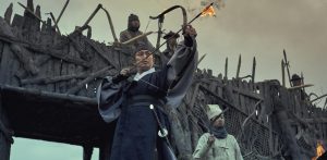 Scène de guerre dans la série Kingdom avec un archer au premier plan, face à une fortification, prêt à tirer une flèche enflammée.