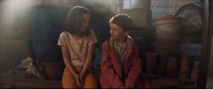 Une petite fille et un petit garçon se sourient intimidés, assis l'un à côté de l'autre dans une pièce entreposant plein de pots de jardin vides, scène du film La dernière vie de Simon.