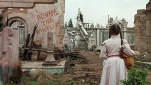 Vue de dos, Dorothy arrive au pays d'Oz, territoire de contes de fée abîmé, en ruines, avec un tag Beware the wheelers sur un des murs.
