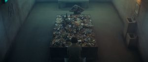 Deux hommes mangent face à face dans une pièce vide et sombre qui ressemble à un hangar, la table est remplie d'aliments en tous genres, scène du film La plateforme.