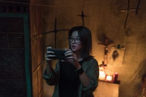 Une jeune femme filme quelque chose avec son smartphone, dans une petite pièce éclairée avec une bougie et des crucifix en bois sur les murs, scène du film Warning : Do not play pour notre concours.