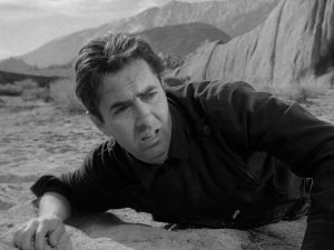 Tyrone Power fatigué à plat ventre sur le sable des plaines, le regard soucieux tourné vers la gauche, scène du film L'attaque de la malle poste.