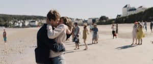Un couple d'adolescents s'embrasse sur une plage, les cheveux au vent, en fond plusieurs badauds, surtout des femmes en robe, scène du film La dernière vie de Simon.