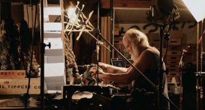 Phil Tippett travaille une maquette, seul assis dans un atelier cerné par des éclairages de cinéma.