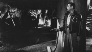 Caché derrière un pan de mur, Humphrey Bogart, l'arme au poing, observe un malfrat à l'arrière-plan, scène du film Tokyo Joe.