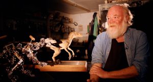 Phil Tippett assis dans son atelier à côté de deux maquettes de tyrannosaures taille réduite.