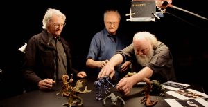 Phil Tippett anime avec ses mains plusieurs maquettes de monstres sur une table noire, devant deux collaborateurs.