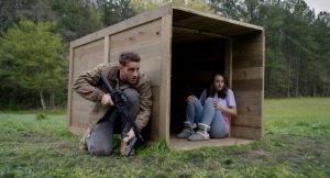 Une jeune femme prostrée dans une caisse en bois, en bordure de fortet, sous la surveillance d'un homme à côté, accroupie, une mitraillette à la main, scène du film The Hunt.