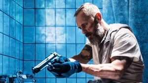 Danny, assis sur le rebord d'une baignoire le revolver à la main dans une salle de bains aux murs à carrelage bleu, scène du film Bluebird.